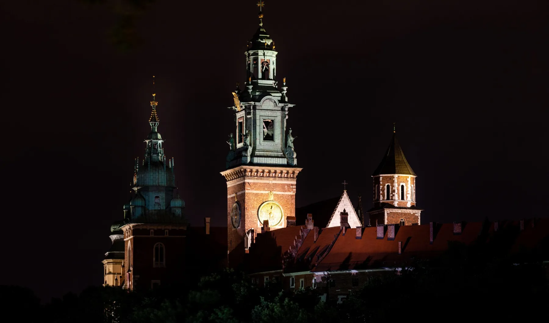 Věže a střechy hradu Wawel zalité světlem reflektorů proti černé noční obloze